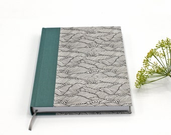 Op de afbeelding: Een notitieboek met harde kaft, een teal stoffen rug en een omslag met een zwart-wit lijnpatroon van gestileerde golven. Het boek is voorzien van een dunne grijze lintbladwijzer aan de onderkant van de rug.