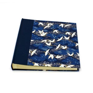 Peut inclure: Un livre relié avec une tranche en tissu bleu foncé et une couverture ornée d'un motif répétitif de grues blanches en vol sur un fond bleu profond, agrémenté de motifs de vagues complexes et de détails floraux subtils dans des tons bleus.
