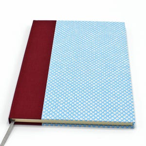 Può includere: Un taccuino con copertina rigida, dorso bordeaux e copertina azzurra con motivo a pois bianchi. È visibile un segnalibro a nastro grigio. Il taccuino è chiuso e appoggiato piatto su una superficie bianca.