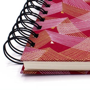 Peut inclure: Un carnet à spirales rouge avec un motif géométrique en or et blanc. Le carnet a une couverture en papier blanc.