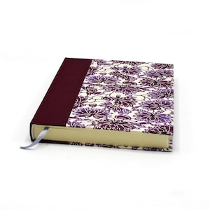 Könnte beinhalten: Ein Notizbuch mit festem Einband, einem dunkelroten Buchrücken und einem cremefarbenen Cover mit violetten und dunkelroten Blumenmustern. Das Buch hat cremefarbene Seiten und ein schmales, hellblaues Lesebändchen an der unteren Kante.