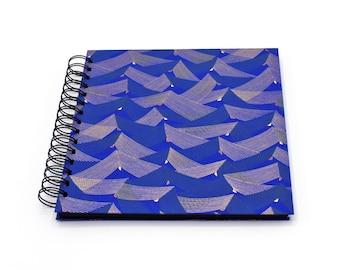 May include: A square spiral-bound notebook featuring a vibrant blue cover with a repeating pattern of stylized, overlapping wave-like shapes in thin, light-colored lines. The binding consists of a black metal wire coil on the left side.