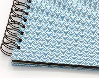 May include: A spiral-bound notebook featuring a repeating pattern of white arched wave shapes on a light blue background. The notebook has a black metal wire coil binding along the left side and a thick black edge along the bottom.
