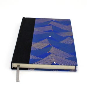 Peut inclure: Un carnet à couverture rigide avec un motif de vagues bleues et dorées. Le dos est noir et un signet en ruban gris est attaché. Le carnet est fermé, avec les pages visibles sur le bord.