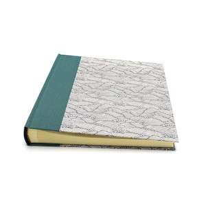 Può includere: Un libro con copertina rigida caratterizzato da un dorso in tessuto color verde acqua e una copertina con un motivo a linee bianche e nere che raffigura forme ondulate ripetitive. Le pagine sono color crema chiaro e ben visibili.