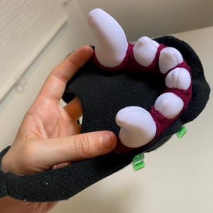 May include: A black plush hand with red and white claws. The hand is attached to a black wrist cuff. The claws are made of fabric and are sewn to the hand. The hand is being held by a person's hand.