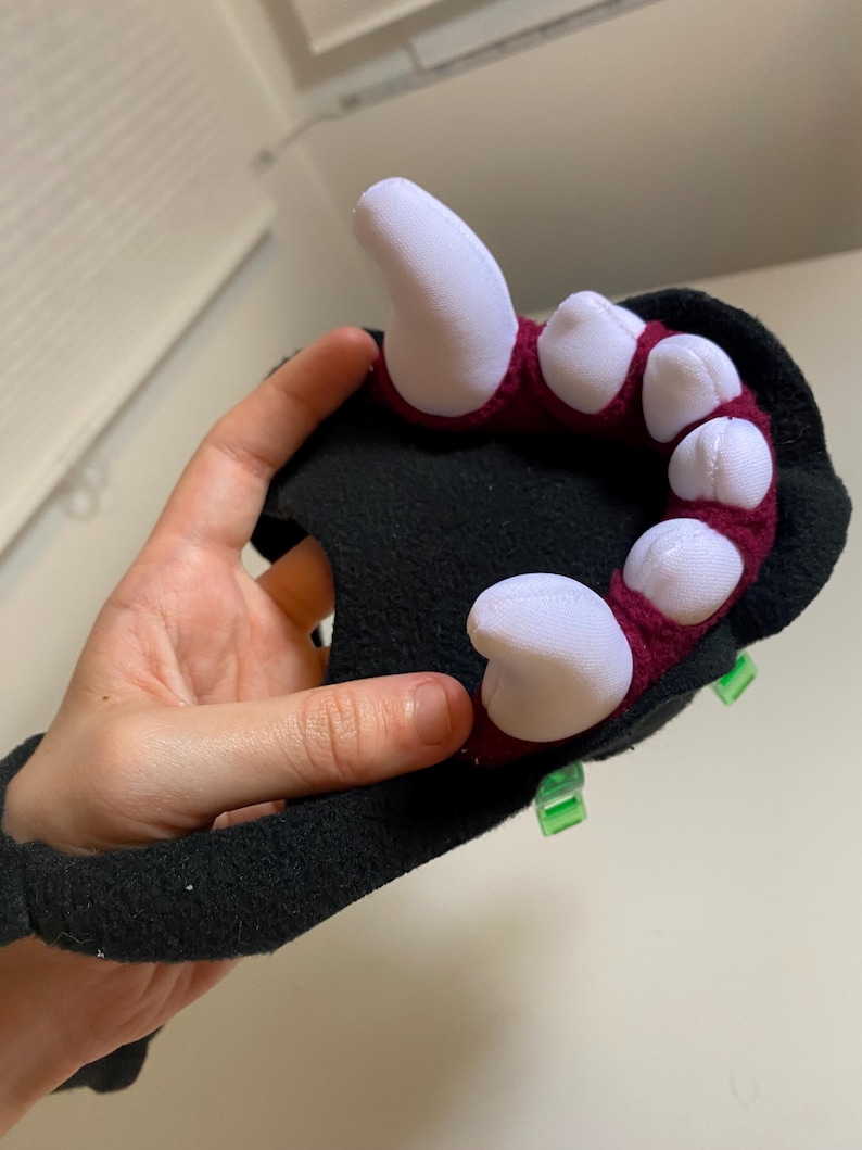 May include: A black plush hand with red and white claws. The hand is attached to a black wrist cuff. The claws are made of fabric and are sewn to the hand. The hand is being held by a person's hand.