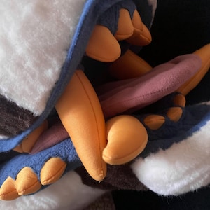 May include: A plush toy with a white and brown fur pattern, blue and orange accents, and a pink tongue sticking out of its mouth.