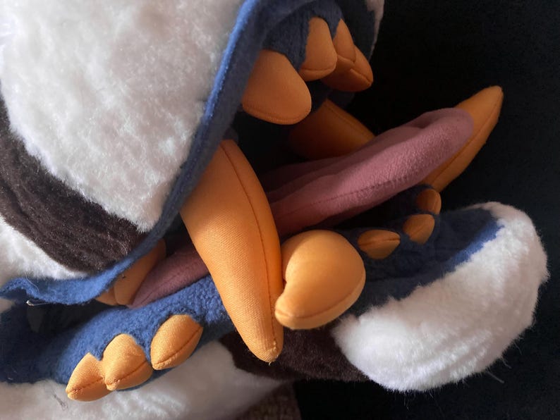 May include: A plush toy with a white and brown fur pattern, blue and orange accents, and a pink tongue sticking out of its mouth.