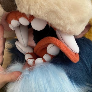 May include: Close-up of a plush animal head with a large open mouth revealing rows of white teeth and a pink tongue. The fur is a mix of light brown, blue, and yellow.