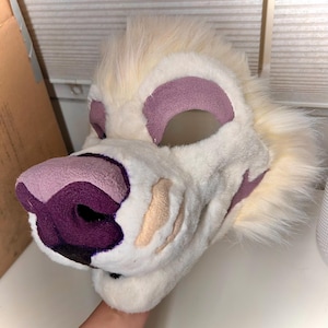 Könnte beinhalten: Ein plüschiger Fursuit-Kopf in Off-White mit lila und beigen Akzenten. Die Schnauze und die Augenpartien sind mit lila Stoff detailliert, und die Ohren sind mit der gleichen Farbe gefüttert. Das Fell ist lang und flauschig.
