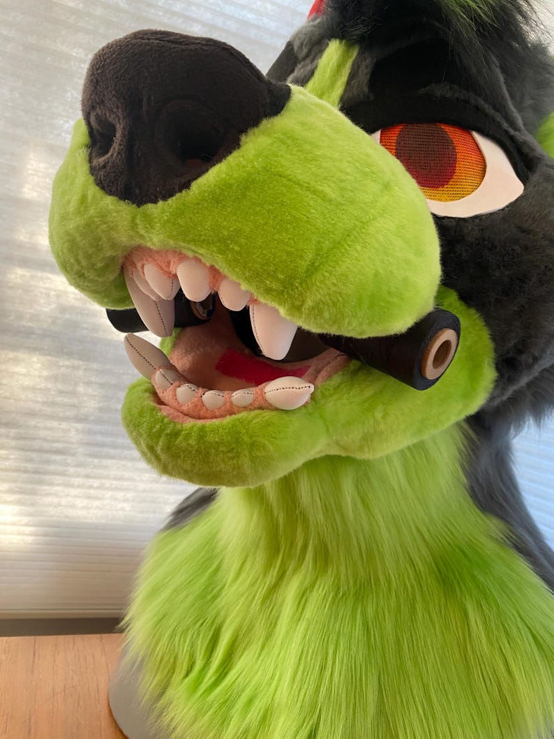 May include: A green and black furry animal head with a large open mouth showing sharp teeth. The head has a red eye with a black mesh pattern.