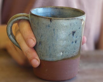 May include: A handmade ceramic mug with a blue-green glaze on the upper portion and a brown base. The mug has a curved handle and is held on a wooden surface. The mug is approximately 4 inches tall.