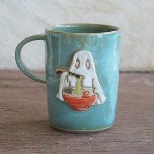 May include: A light blue ceramic mug featuring a raised, sculpted white ghost figure eating from a small orange bowl with chopsticks. The mug has a smooth, glossy finish and a curved handle on the left side, showcasing a detailed relief design.