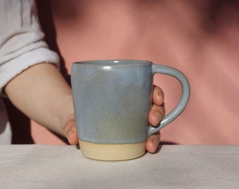 May include: A light blue ceramic mug with a cream-colored rim and handle. The mug is sitting on a light gray and white striped tablecloth.