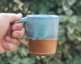 May include: A hand holding a blue and brown ceramic mug. The mug has a light blue glaze with a brown band around the bottom.