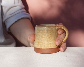 May include: A hand holding a small, light brown ceramic mug with a brown rim. The mug is sitting on a white linen tablecloth.