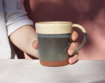 May include: A handmade ceramic mug with a unique design. The mug features a beige rim, a dark blue band, and a dark green body, with a brown base. The handle is a mix of blue and green. The mug is held in a person's hand.