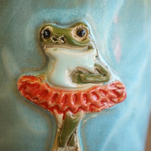May include: A light blue ceramic mug featuring a raised, three-dimensional sculpted green frog wearing a bright orange ruffled tutu. The frog is positioned in the centre of the mug, with its arms crossed and legs crossed at the bottom.