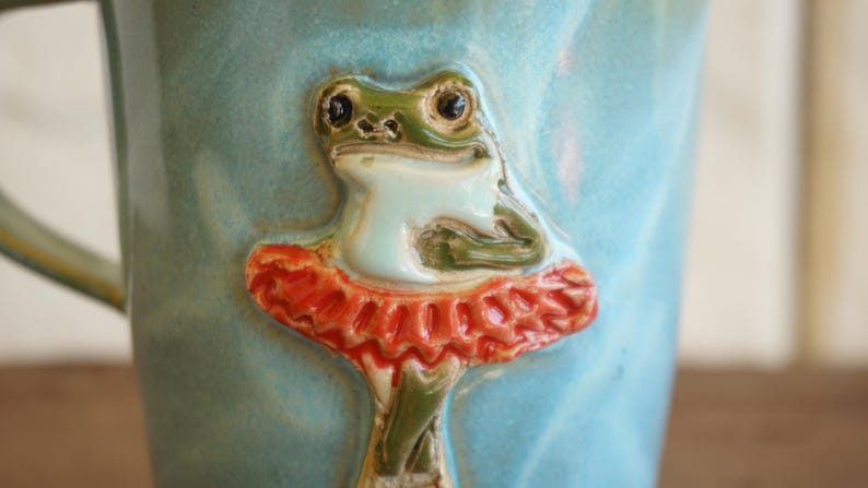 May include: A light blue ceramic mug featuring a raised, three-dimensional sculpted green frog wearing a bright orange ruffled tutu. The frog is positioned in the centre of the mug, with its arms crossed and legs crossed at the bottom.