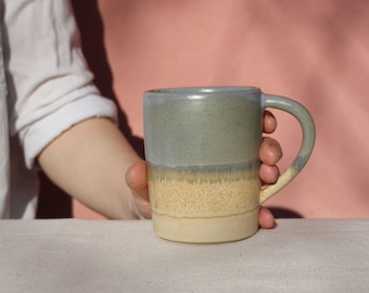 May include: A hand holds a ceramic mug with a light blue, gray, and yellow glaze. The mug has a rounded handle.