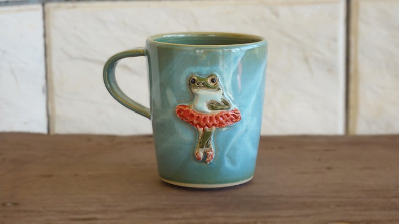 May include: A light blue ceramic mug featuring a raised, three-dimensional sculpted frog wearing a red tutu. The mug has a smooth, glossy finish with subtle variations in color and a single handle on the left side of the cylindrical body.