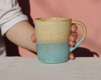 May include: A ceramic mug with a speckled, light brown glaze on the top half and a speckled, light blue glaze on the bottom half. The mug has a rounded handle.