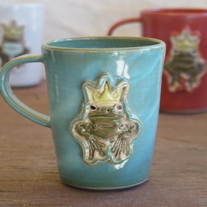 May include: A light blue ceramic mug featuring a raised, sculpted green frog wearing a yellow crown on its front. Two additional mugs, one white and one red, each with a similar frog design, are positioned in the blurred background.