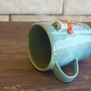 May include: A light blue ceramic mug featuring a three-dimensional sculpted frog figure wearing a textured orange scarf attached to the exterior surface. The mug has a smooth glazed finish, a curved handle, and a wide circular opening at the top.