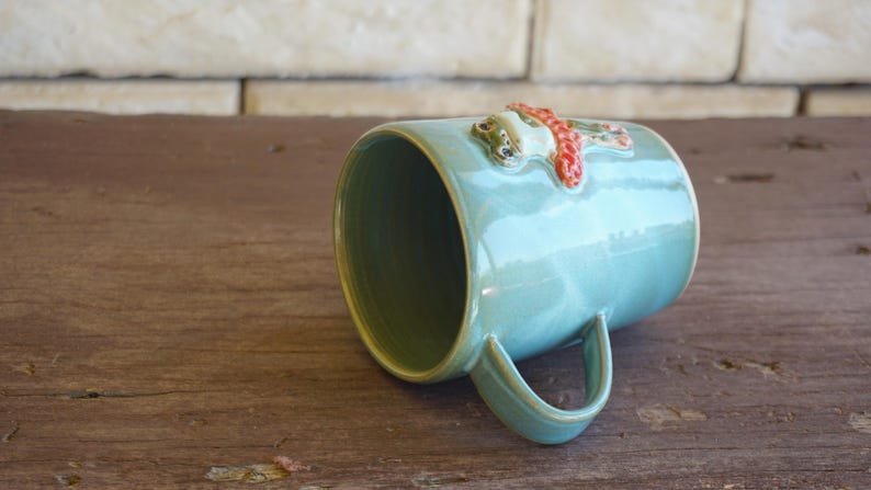 May include: A light blue ceramic mug featuring a three-dimensional sculpted frog figure wearing a textured orange scarf attached to the exterior surface. The mug has a smooth glazed finish, a curved handle, and a wide circular opening at the top.