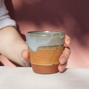 May include: A hand holds a ceramic mug with a light blue glaze on the top half and a brown glaze on the bottom half.