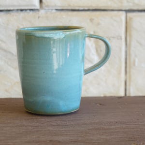 May include: A ceramic mug with a smooth, glossy finish in a light turquoise blue colour. The mug features a cylindrical body that tapers slightly towards the base, a rounded rim, and a single curved handle attached to the side of the vessel.