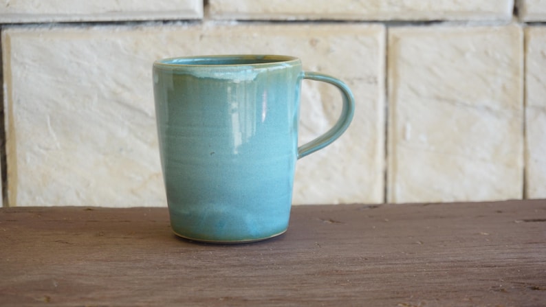 May include: A ceramic mug with a smooth, glossy finish in a light turquoise blue colour. The mug features a cylindrical body that tapers slightly towards the base, a rounded rim, and a single curved handle attached to the side of the vessel.
