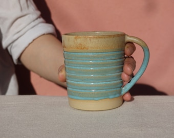 May include: A hand holds a light blue and beige ceramic mug with a striped design. The mug has a rounded handle.