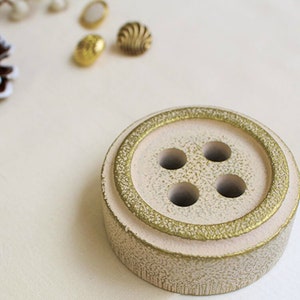May include: A white ceramic button-shaped trinket dish with a gold rim and four holes in the top. The dish is perfect for holding small items like jewelry or keys.
