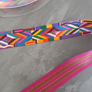 Op de afbeelding: Een kleurrijk lint met een geometrisch patroon in tinten rood, oranje, geel, groen, blauw, paars en wit. Het lint ligt op een grijs oppervlak.