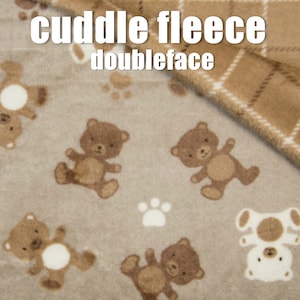 Pode incluir: Um tecido de lã marrom e branco com um padrão de urso de pelúcia e pegada de pata. O tecido é de dupla face, com um lado com um padrão xadrez marrom e branco. O tecido é etiquetado como "cuddle fleece doubleface".