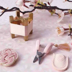 May include: A selection of small, decorative items in pink and gold. Includes a miniature gold and pink stand, a woven pink coaster, pink scissors, and a pink leather sheath. Cherry blossom branches are in the background.