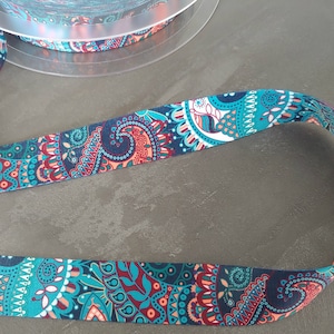 Peut inclure: Bobine de ruban à motifs avec un motif répétitif en turquoise, marine et rouge. Le ruban présente un motif de type cachemire avec des éléments floraux et géométriques. Le ruban est sur une bobine en plastique transparent.