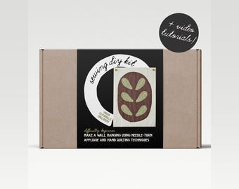 Puede incluir: Una caja de cartón marrón con un círculo blanco y el texto "sewing diy kit" en la parte delantera. La caja contiene un kit para hacer una pared colgante con un diseño marrón y verde. El texto "video tutorials included" también está en la caja.