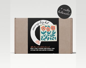 Puede incluir: Una caja de cartón marrón con una etiqueta negra que dice "Sewing DIY Kit" y presenta un diseño floral colorido. La etiqueta también incluye el texto "Vídeos tutoriales incluidos" y "Dificultad: Intermedio/Avanzado". La caja incluye instrucciones para hacer una pared colgante usando la técnica de aplicación de aguja vuelta y técnicas de acolchado a mano.