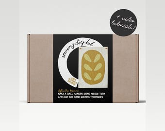 Puede incluir: Una caja de cartón marrón con una etiqueta negra que dice "Sewing DIY Kit" e incluye una imagen de una hogaza de pan. La etiqueta también dice "Vídeos tutoriales incluidos" y "Dificultad: Principiante". La caja es para un proyecto de aplique de vuelta de aguja y acolchado a mano.