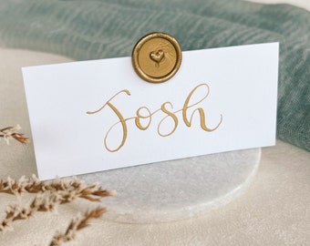 Può includere: Un segnaposto bianco con il nome "Josh" scritto in calligrafia dorata. Un sigillo di cera dorato con un motivo a forma di cuore è attaccato alla parte superiore della carta.