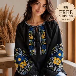 Puede incluir: Una blusa negra de manga larga con intrincados bordados de punto de cruz en patrones florales amarillos, azules y verdes. La prenda incluye un cuello redondo con un cierre de lazo terminado con borlas negras y puños en las mangas.