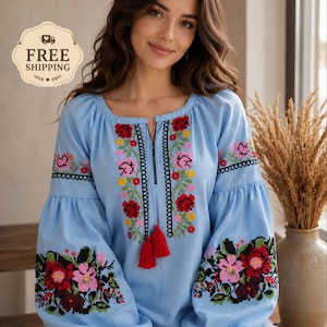 Peut inclure: Un chemisier bleu clair à manches longues avec une encolure carrée et une fente centrale dotée de pompons rouges. Le vêtement est orné de broderies au point de croix représentant des motifs floraux rouges, roses, jaunes et verts sur le buste.