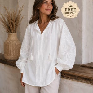 May include: A white long-sleeved blouse featuring a V-neckline with two hanging tassels, vertical pleats down the center front, and intricate floral embroidery on the voluminous sleeves. The blouse is paired with light-colored trousers.