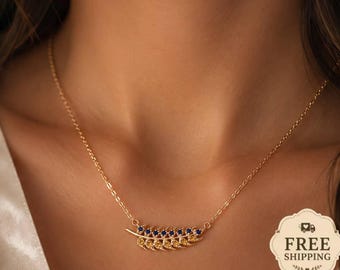 May include: A gold-toned necklace featuring a delicate horizontal feather-shaped pendant. The pendant is embellished with a row of small, round, deep blue gemstones set along the upper edge of the gold feather structure, suspended on a fine chain.
