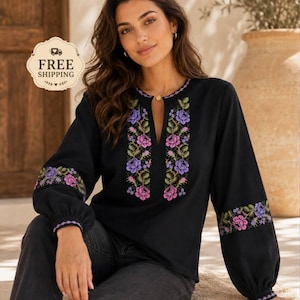 Op de afbeelding: Een zwarte blouse met lange mouwen, een split-halslijn en gedetailleerd bloemenborduursel in paarse, roze en groene tinten. De borduurpatronen zijn verticaal op de borst en horizontaal over de mouwen geplaatst.