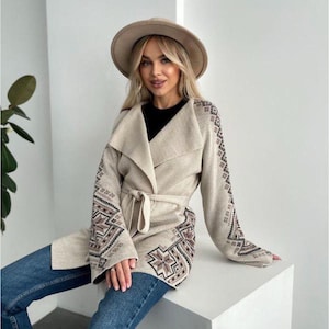 Peut inclure: Un cardigan en tricot beige avec un col châle et une ceinture à nouer. Le cardigan présente un motif géométrique sur les manches et une bordure brune contrastante. Le cardigan est porté sur un haut noir et un jean bleu.