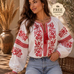 Op de afbeelding: Een witte blouse met lange mouwen voorzien van ingewikkeld rood bloemenborduursel op de voorzijde en mouwen, een ronde halslijn met een centrale split en rode kwastjes, gecombineerd met een lichtblauwe spijkerbroek.